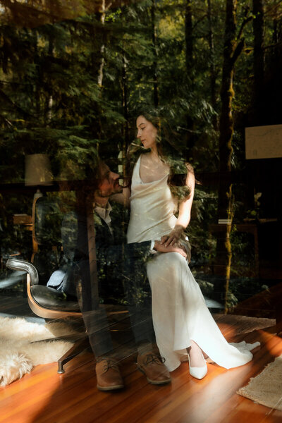Portrait of bride and groom through a window into a cabin with the reflection of the Bend, Oregon trees surrounding them