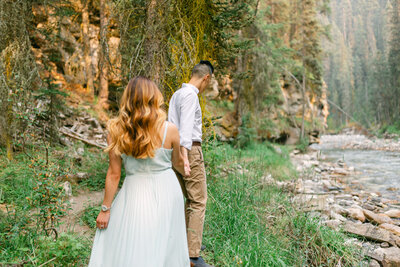 This vibrant photograph captures a couple walking along the scenic Johnston Canyon in Banff National Park during their summer engagement shoot. Surrounded by lush greenery and cascading waterfalls, the moment reflects the natural beauty and joy of their adventure together.