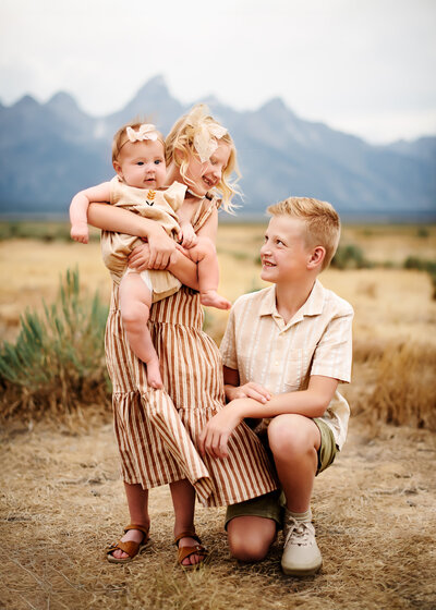 Three siblings standing together in their cute clothing and smiling at one another. 