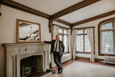 Groom standing for top wedding portraits at Elsie Perrin Williams estate in front of stone fireplace with his forearm resting on the tall mantle and legs casually crossed. Groom is looking over his shoulder off in the distance.