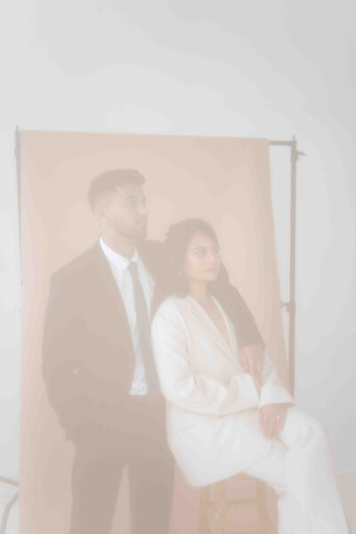 Blurry photo of couple lean on to each other while looking forward  in front of tan photo backdrop