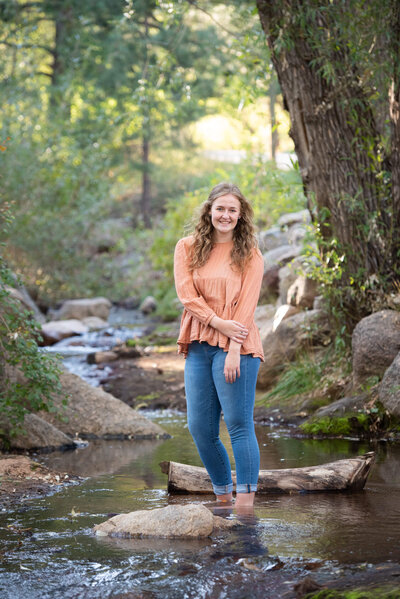 A woman holds her arm while standing in a shallow creek in jeans and pink shirt