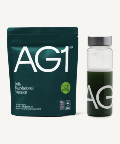 A green pouch of AG1 daily nutrition powder next to a glass bottle filled with a dark green liquid and labeled “AG.” This health supplement is marketed for energy, gut health, and overall wellness.
