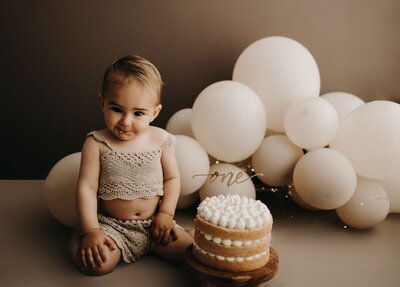 Little one in beige knitted outfit sitting on soft carpet, gazing sweetly with neutral backdrop and soft tones.