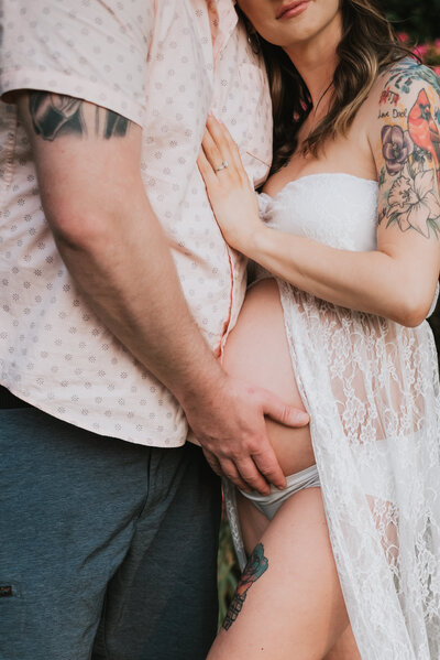 A close up photo of dad holding moms pregnant belly in Baltimore Maryland while mom is wearing a white lace dress and dad is wearing a pink shirt and blue shorts.