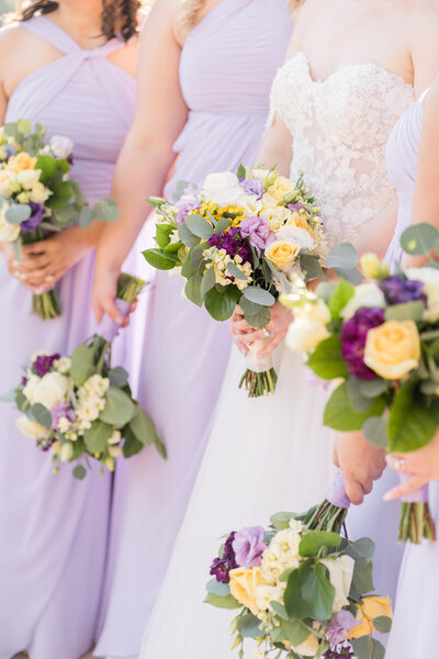 a bride and her bridesmaids hold their bouquets of white, purple, and yellow florals