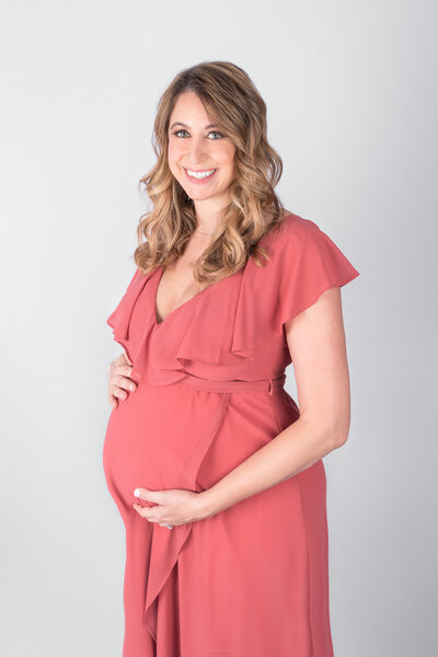 A pregnant woman wearing a pink dress holds her baby bump while smiling and standing in front of a grey backdrop
