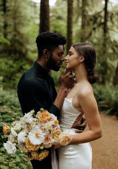 Bride and groom embracing in a forest setting, holding a bouquet of pastel flowers.