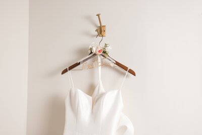 A modern, elegant white wedding dress hangs on a custom wooden hanger with “Mrs.” written in gold wire. A soft pink flower and white floral detail adorn the top of the hanger, creating a delicate, romantic touch against a simple beige wall. This classic bridal detail was photographed by Heartful Films, a Jacksonville wedding photographer and videographer known for capturing the beauty in both timeless details and emotional storytelling.