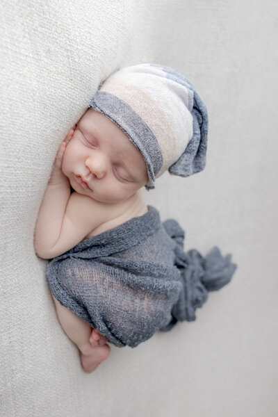 Lehigh Valley Newborn Photographer newborn boy on a cream blanket with his hands under his head and a blue wrap over him wearing a blue cream and white hat