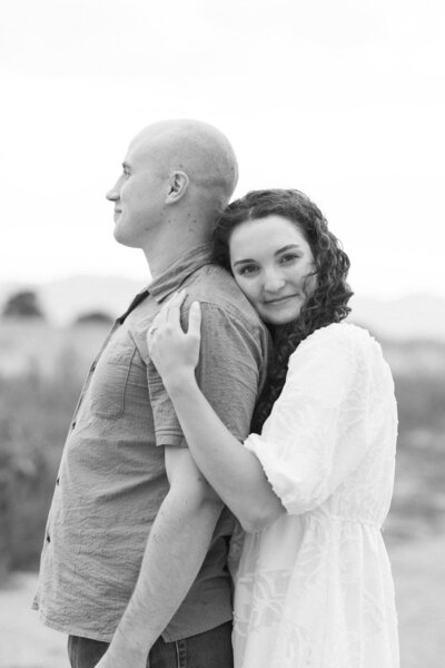 A woman stands behind a man and wraps her arms around his. She looks at the camera while he looks forward.