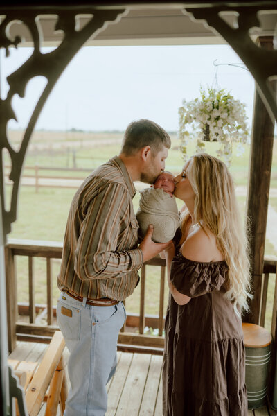 couple kissing their newborn baby on their front porc