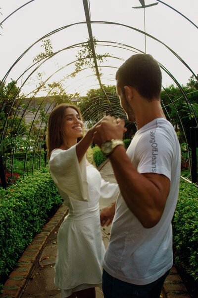 A couple dances joyfully under an arched trellis in a lush garden. The woman wears a white dress, smiling, while the man in a white t-shirt leads. Romantic ambiance.