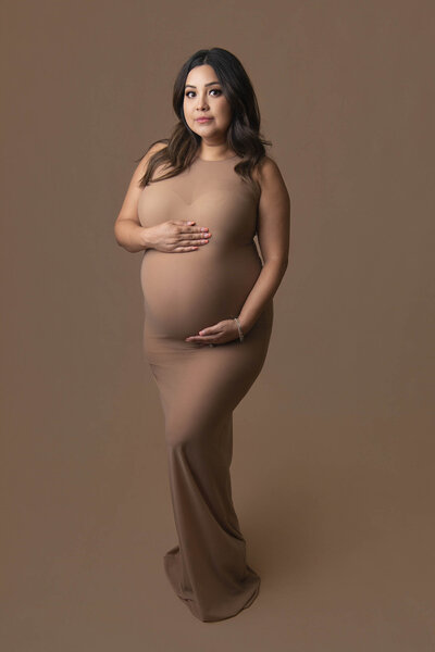 A mother to be stands in a studio smiling down to her bump in a white tule maternity gown