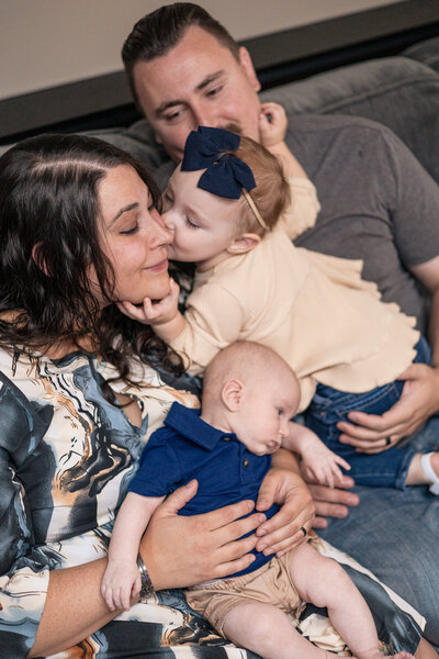 Family of 4 on the couch with their toddler daughter kissing mom on the cheek.