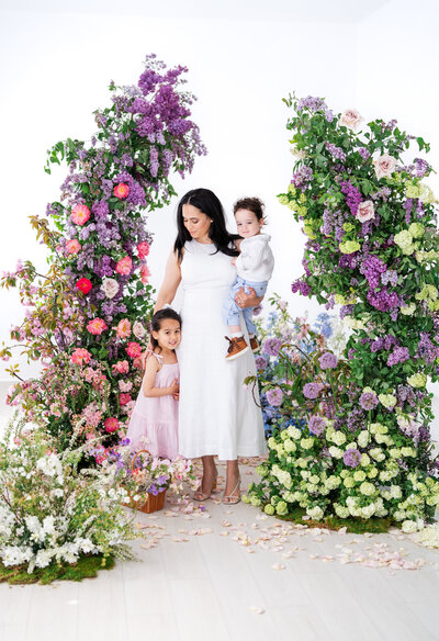 A pregnant woman in a flowy white and blue floral dress sits on a chair, smiling as a young boy hugs her belly, surrounded by lush floral arches of purple, pink, and green blooms.