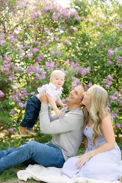 a family sits in front of lilac bushes, and the dad puts the smiling baby up into the air and both mom and dad smile up at him.