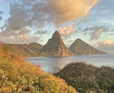 A vibrant sunset over the Pitons in Saint Lucia, with the iconic twin peaks rising from the ocean. The foreground features lush green foliage, illuminated by the warm, golden light of the setting sun.