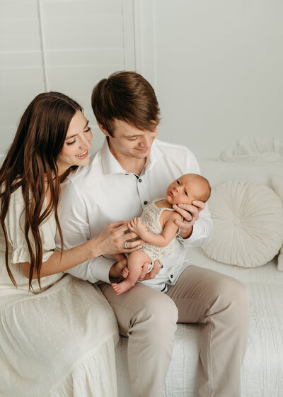 Dad is holding his newborn baby in her arm while both him and the mother is looking down at their baby and smiling . Photo  taken by Lisa Lefevre in Atlanta