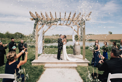 Confetti showers over the bride and groom as they kiss during their outdoor ceremony