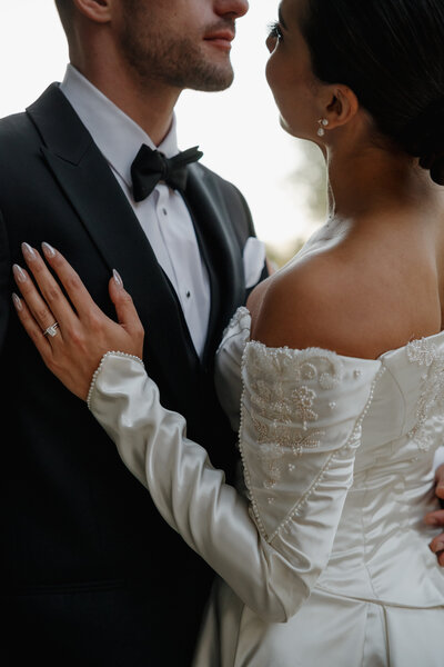 Bride in a beaded off-the-shoulder wedding dress rests her hand on the groom’s chest, showing her ring and delicate manicure.