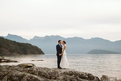 A couple standing facing each other on a rock in front of the Ocean and Mountains at Camp Fircom on Gambier Island.
