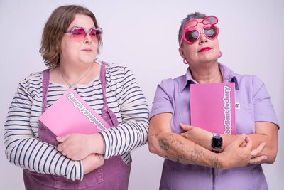 Two people pose side by side against a pale studio backdrop, each hugging a bright pink notebook. Left is Lucy in a striped top with purple overalls and rose-tinted sunglasses. Right is Pippa in lilac dungarees with round pink heart-shaped sunglasses and tattooed forearms, both striking a playful, mock-serious stance.