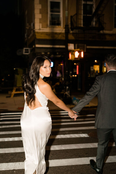wedding couple holding hands and walking across the street while the bride looks over her shoulder