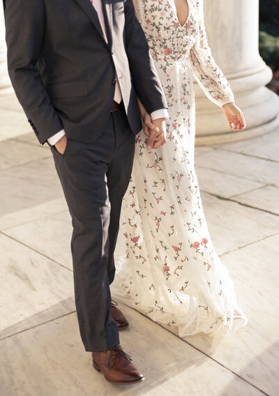 A couple holds hands while walking. The person on the left is in a dark suit and light tie; the person on the right is wearing a long white dress with floral patterns. They are walking on a stone surface with white columns in the background.