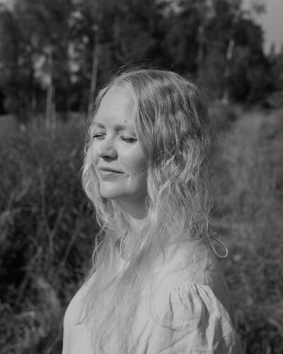 Henkilökuva naisesta, joka seisoo silmät kiinni kasvot aurinkoa kohti ja hymyilee maaseudulla Suomessa