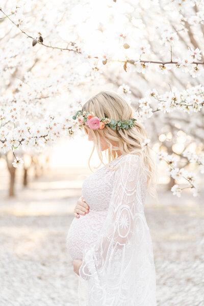 A maternity session by Bay area photographer shows a mother holding her baby bump dressed in a lace white gown with a rose flower crown standing in a field of almond blossoms.