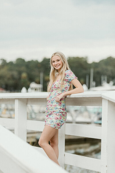 Senior albrey leans against a white wooden rail at the Isle of Hope Marina during her senior session. She is wearing a short fitted floral dress and props her elbow up on the bannister and smiles with knee bent slightly.