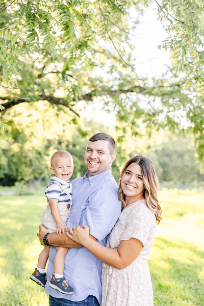 Mom hugging dad from behind while dad holds baby boy and they all smile at the camera in a grassy field on a bright summer evening in Ohio.