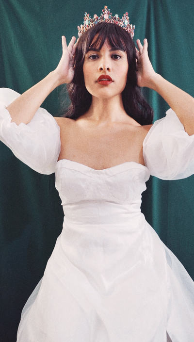 Founder of Elyana Studio, Elysia Anais, is standing in a white ballgown dress, holding a crown on top of her head against a dark green background for Elyana Studio Showit Website branding