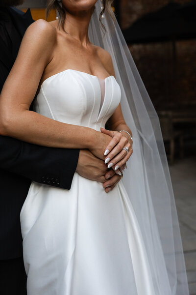Bride and groom holding each other, gazing out of a window at their wedding venue, captured in soft, natural light for a romantic and intimate moment.