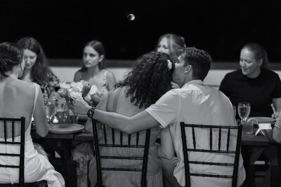 A person with their arm around their partner chair as they lean into each other 