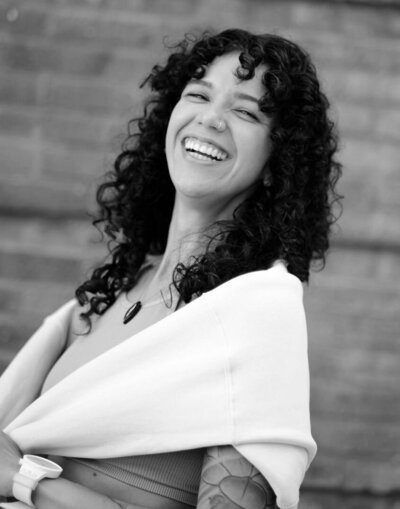 Black and white photo of woman with long, curly dark hair smiling at the camera while holding a white sweater wrapped around her shoulders.