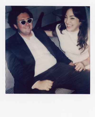 A couple sits on a couch, laughing, on their wedding day.