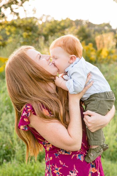 A white woman with red hair is holding her infant, who is playfully biting her chin