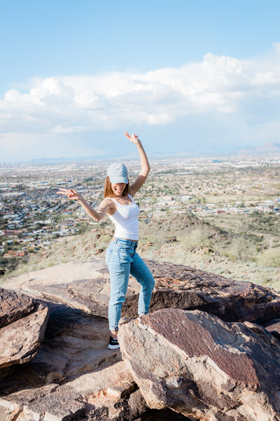 Picture of a girl posing on top of a mountain with peace signs. She is wearing a baseball hat.