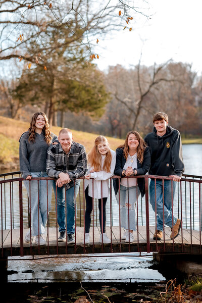 a family of 5 is leaning on the railing of a bridge in Susan park. The lake is under the bridge and behind them.