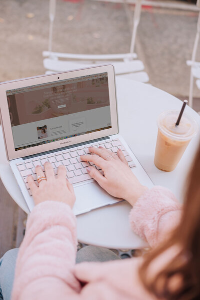Close-up of Madison’s hands typing on a MacBook Air with a pink keyboard cover, sitting outside at a café table.