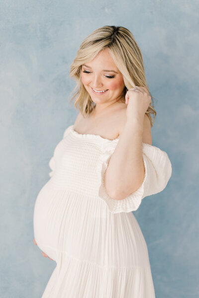 A pregnant woman wearing a pink dress stands in a field holding her belly.