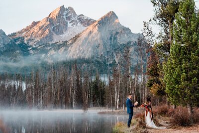 A bride and groom say their vows to each other at the foot of a mist covered lake with jagged mountains rising up behind them, touched with snow.  They are on their 2 day adventure luxe elopement in the mountains in Idaho. 