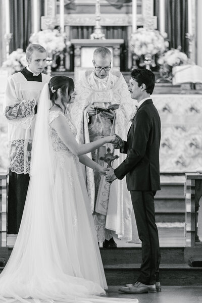 Black and white photo of bride and groom holding hands while standing in front of priest in church