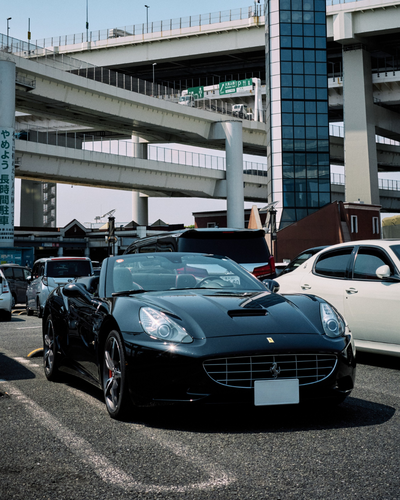 Front view of a black Ferrari California convertible parked under Tokyo expressway ramps at the iconic Daikoku Parking Area—part of the Tokyo Luxe Drive fleet.