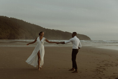 Husband spins his wife around on a beach on the Oregon Coast, her white dress blowing in the wind.