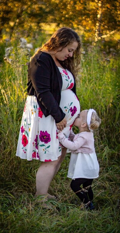 a little girl kisses her moms pregnant belly in a field in Edmonton 