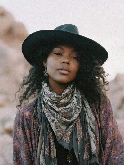 A person with curly hair wears a wide-brimmed black hat, patterned scarf, and paisley shawl, standing outdoors with a neutral expression—embodying the creative flair of Sweet Pea Ink Creative’s brand design.
