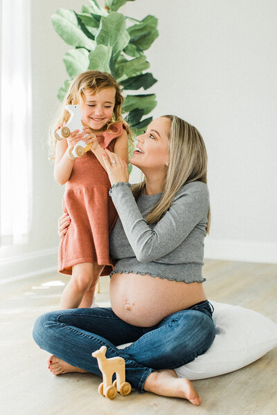 pregnant mom sits of the floor with crossed legs and older daughter hugs her and laughs